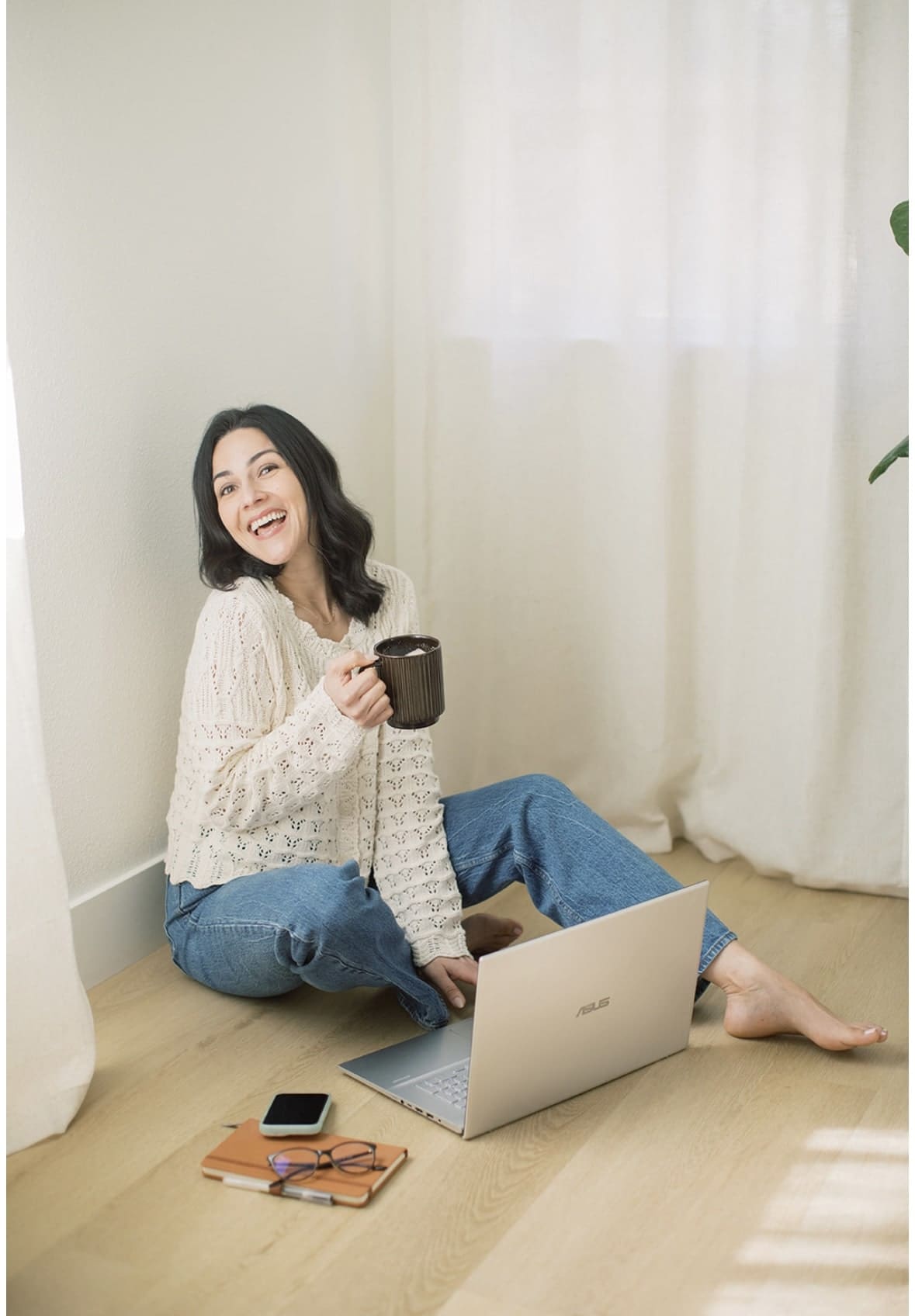 Gwen sitting on the ground in a white knit top and jeans, holding a cup of coffee and looking to the side, a silver laptop lays open in front of her along with a phone, leather notebook and eyeglasses